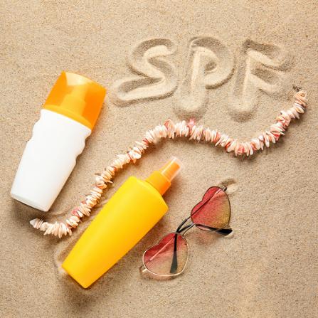Deux crèmes solaires sont posées sur le sable à côté d'une paire de lunettes de soleil en forme de cœur et le terme SPF est inscrit dans le sable