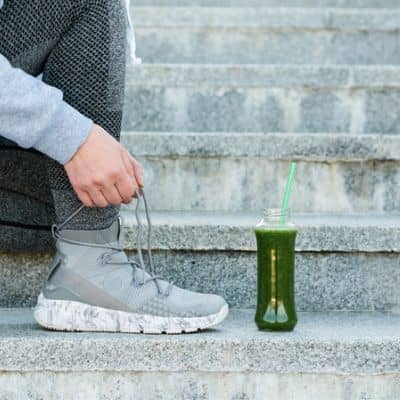 femme qui refait ses lacets de chaussures de courses à pieds avec un jus détox à côté