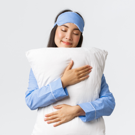Une femme souriante tient sous oreiller dans les bras et porte un masque de nuit remonté sur son front