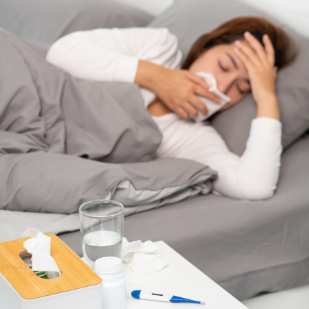 Une femme dans son lit tient un mouchoir dans une main et pose l'autre sur son front, un thermomètre et une boîte de mouchoirs sont posés sur la table à côté
